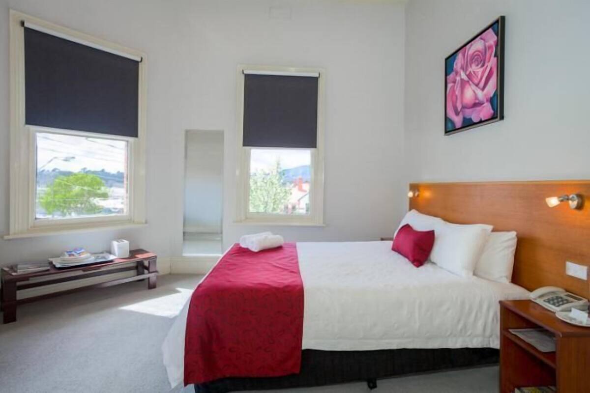 Ein Hotelzimmer mit Bett, Nachttisch mit Telefon, Fenster mit Blick und einem Bild an der Wand.