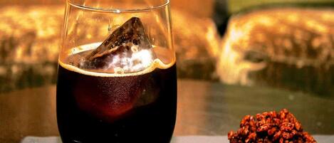 Un vaso de bebida oscura con hielo y un objeto de color oscuro, posiblemente una fruta, sobre el borde. Un montón de objetos pequeños, redondos y marrones se ve sobre la mesa junto al vaso.