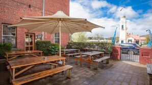 Een terras buiten met houten tafels en banken, een grote parasol en een bakstenen gebouw op de achtergrond.