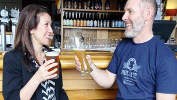 Dos personas sostienen vasos de cerveza en un bar con estantes llenos de botellas y un letrero que dice