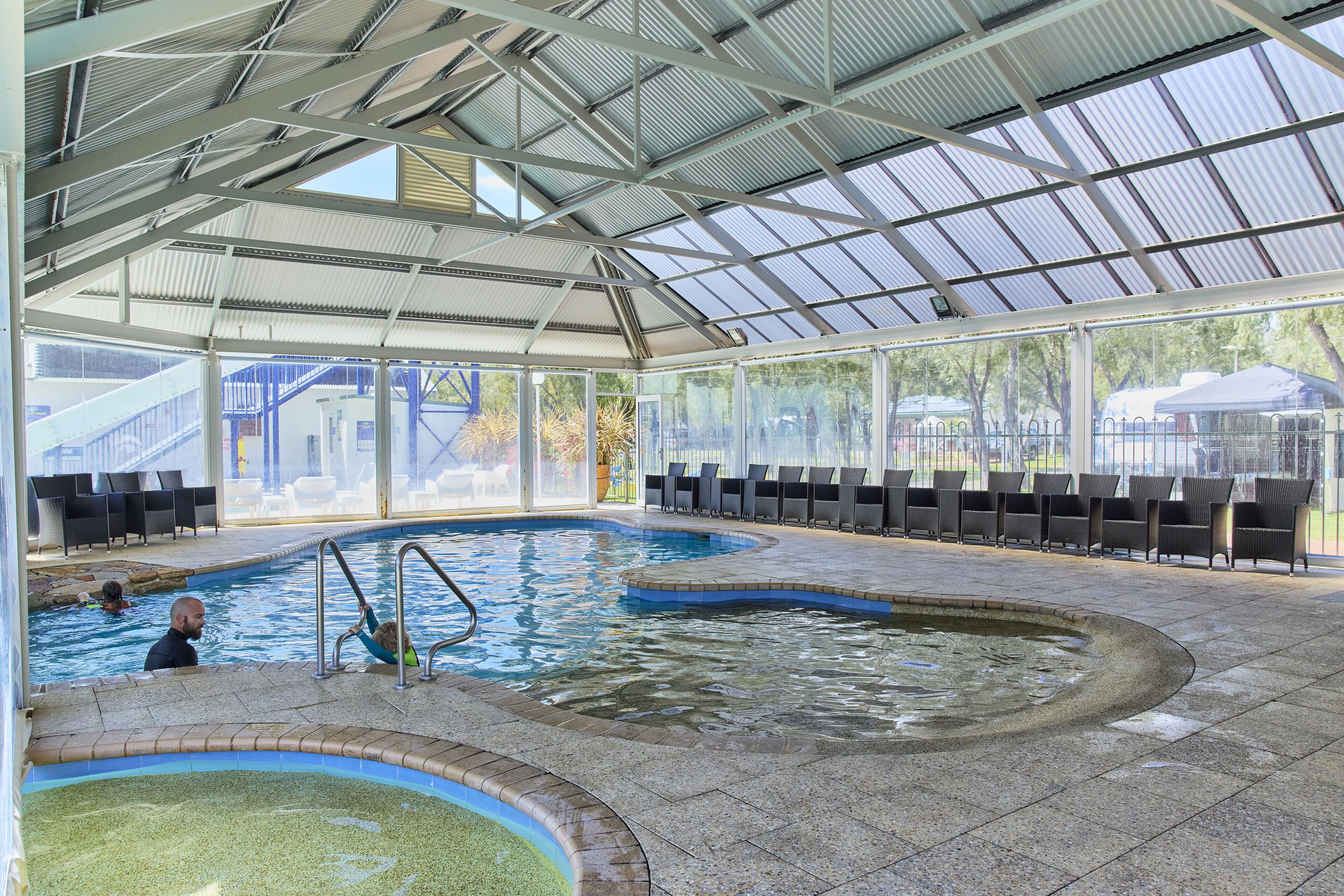 An indoor swimming pool with clear glass walls, a covered roof, and multiple seating areas.