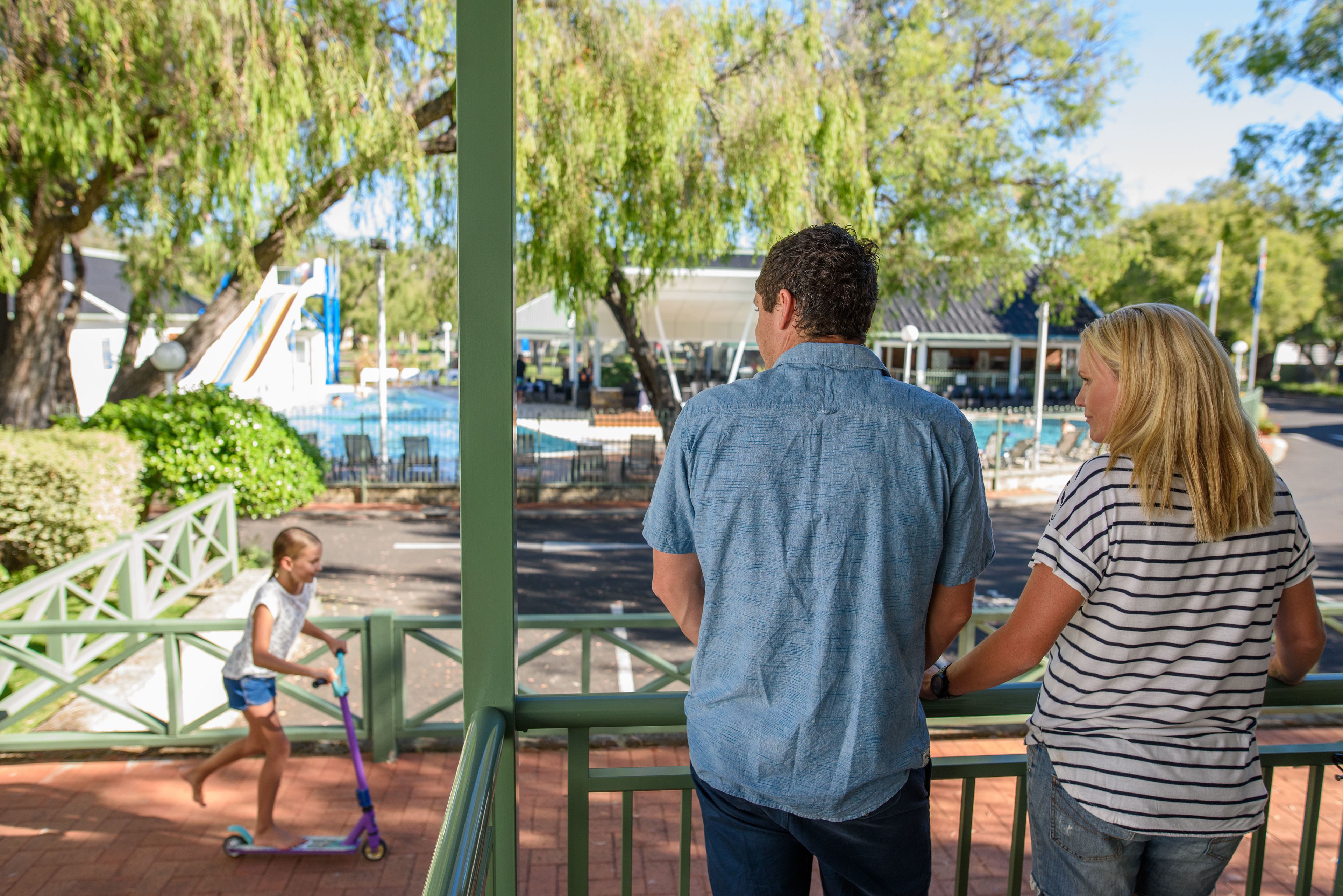 A man and a woman standing by a railing, overlooking a playground with a slide and a child on a scooter.