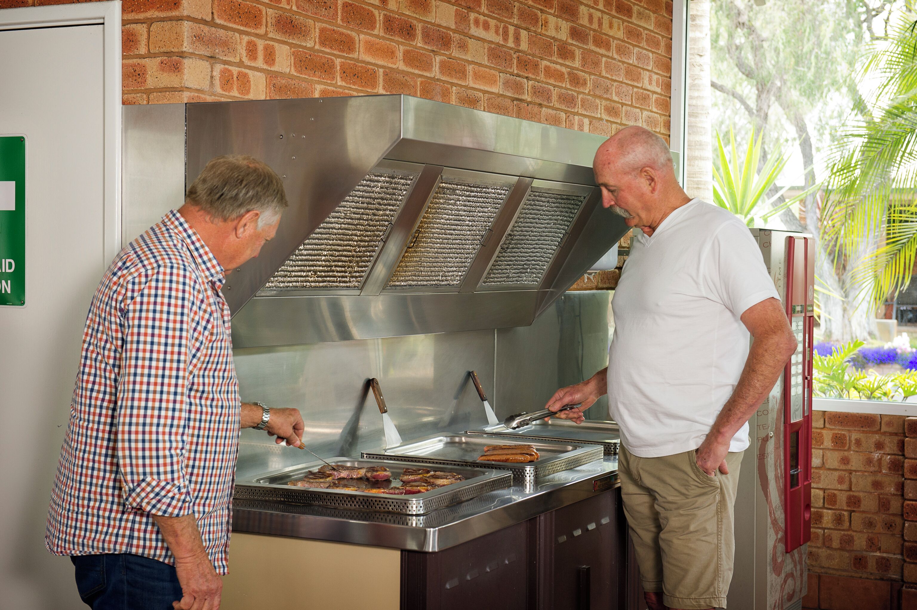 Two men at a food service counter with a brick wall and a window with plants.