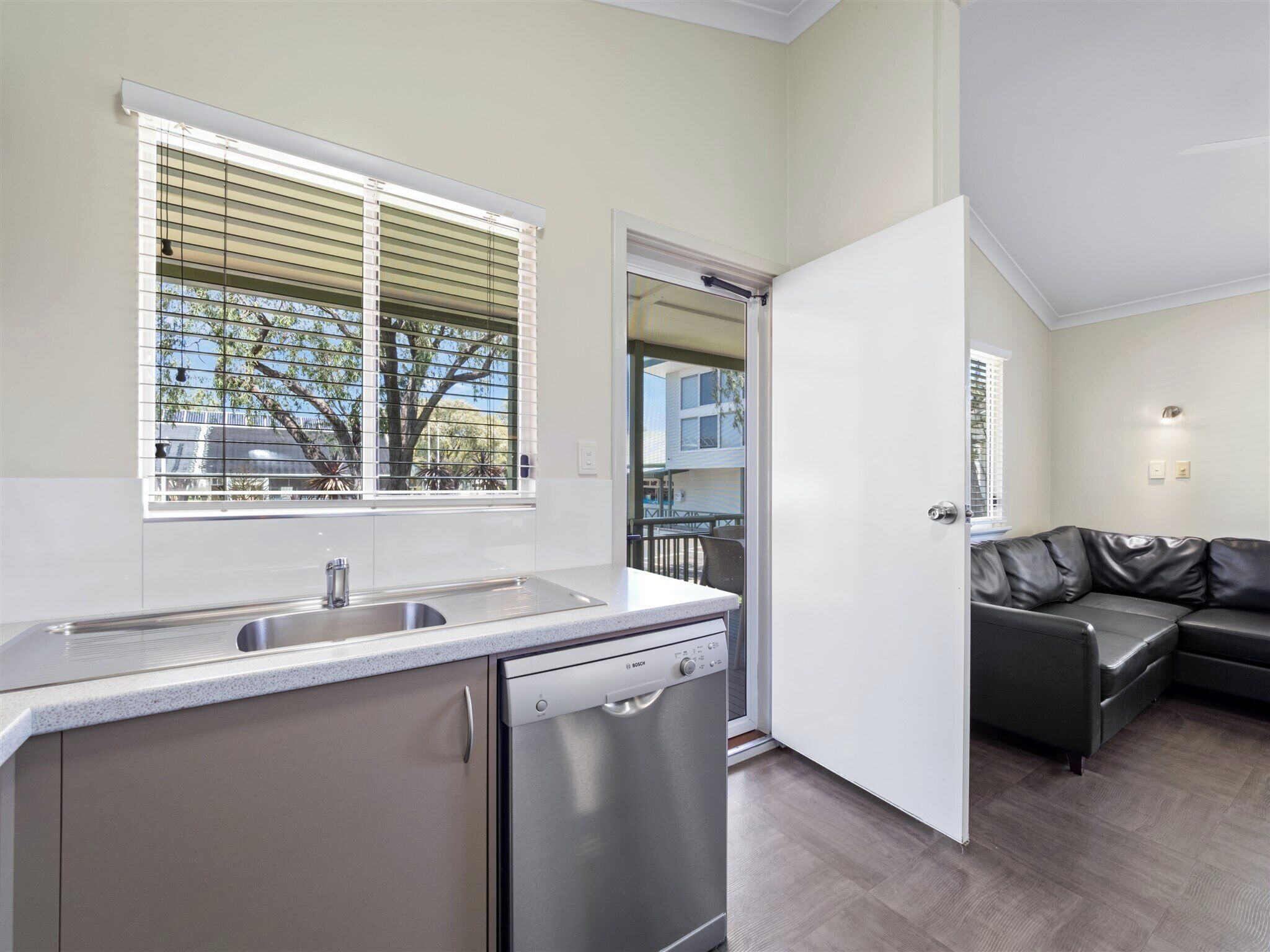Cocina compacta con lavavajillas, ventana con persianas y vista a un balcón.