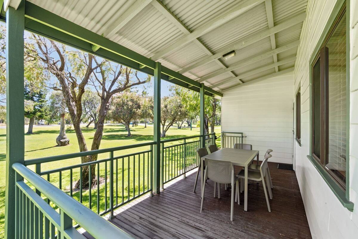 A covered porch with a table and chairs, a view of a grassy park with trees, and a white building with green trim.