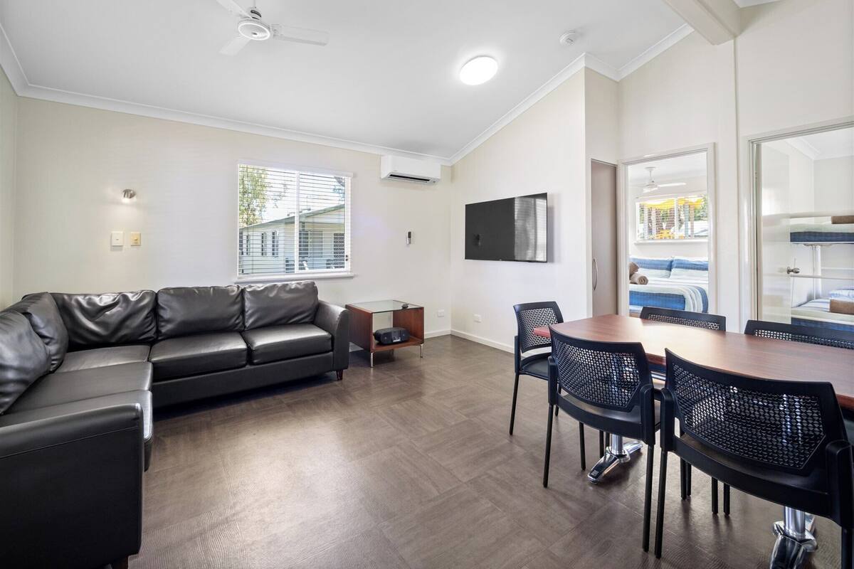 A modern living room with a black leather sofa, a dining table with four chairs, a ceiling fan, and a mounted TV.