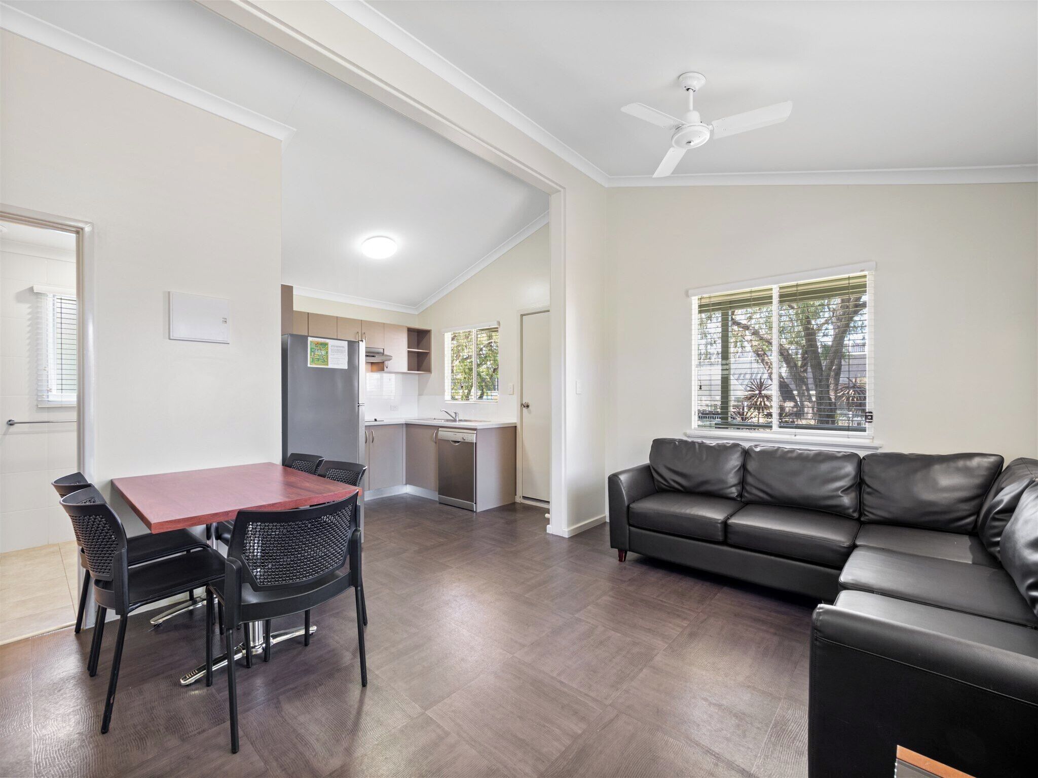 A modern living room with a black leather sofa, a dining table with four chairs, a kitchen area with a refrigerator and sink, and a ceiling fan.