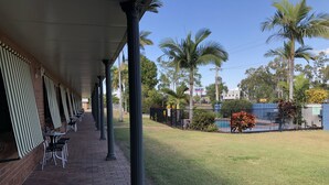 A covered outdoor seating area with brick flooring and metal chairs, palm trees, and a fenced pool area. - True Blue Motor Inn (Parkhurst)
