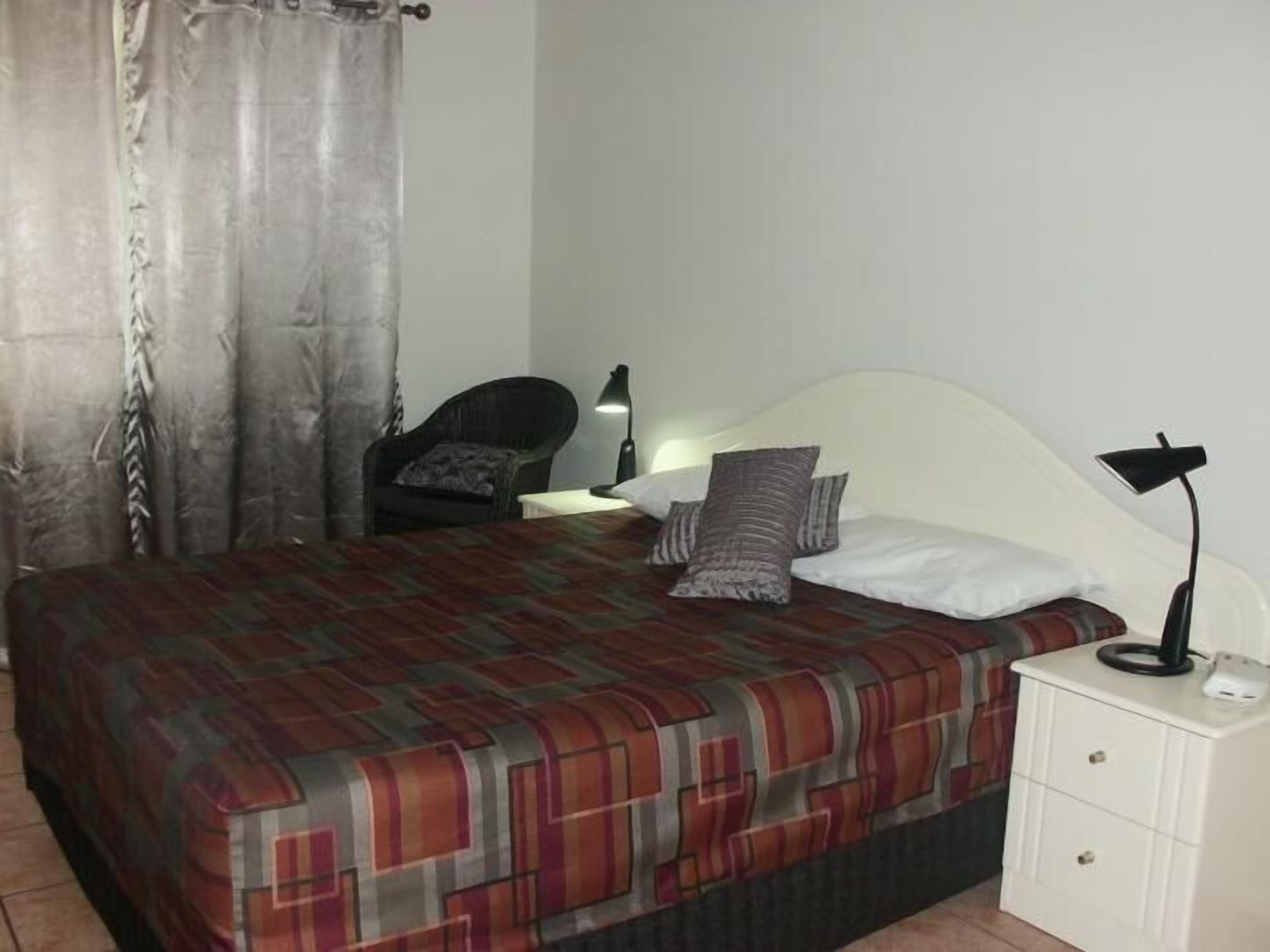 A bed with a patterned bedspread, a bedside lamp, a chair, and a nightstand with a lamp.