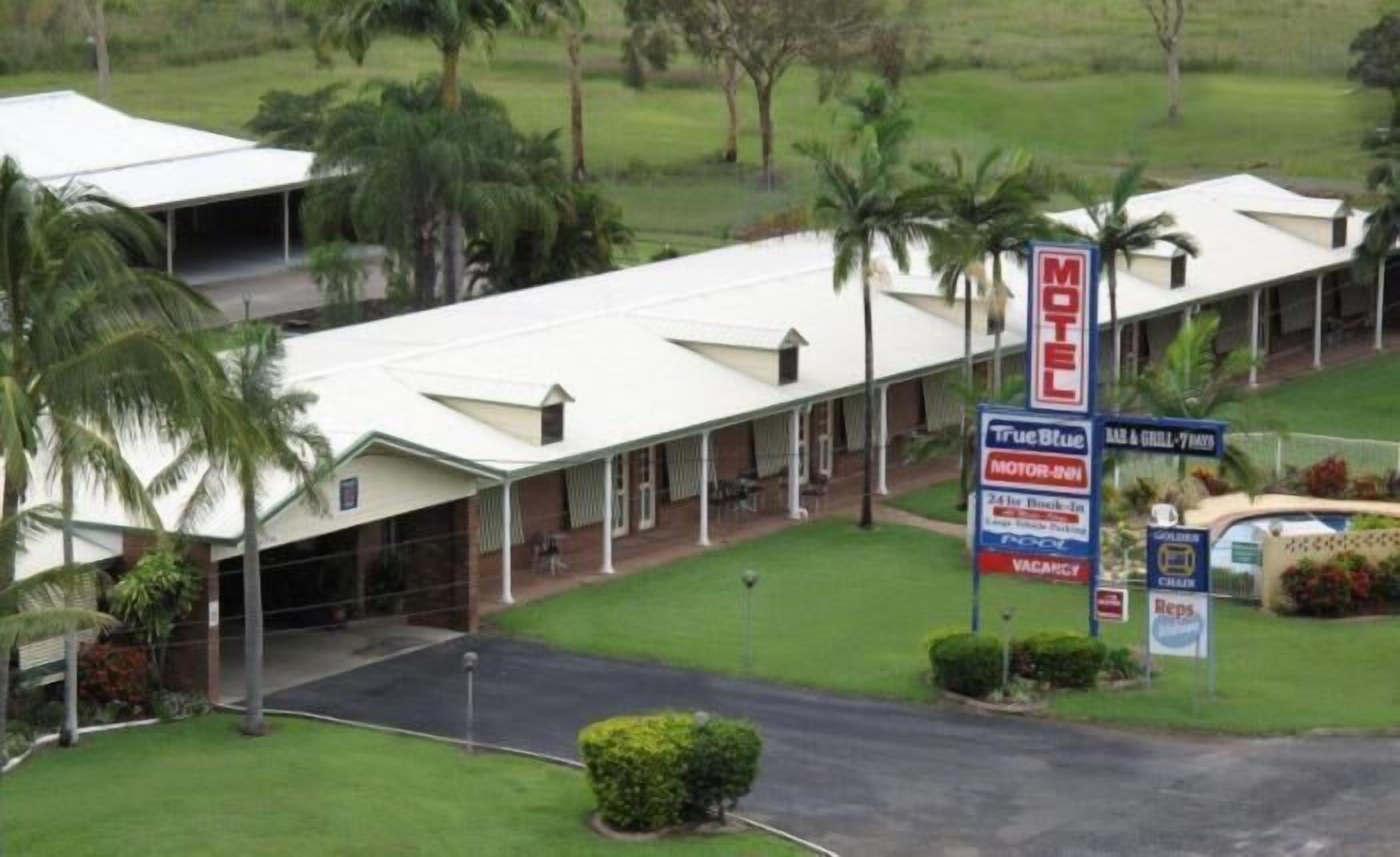 A motel with a covered parking area, a signboard advertising True Blue Motor Inn, and a view of the surrounding greenery.
