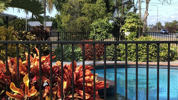 A swimming pool surrounded by a metal fence, with various plants and trees in the background. - True Blue Motor Inn (Parkhurst)