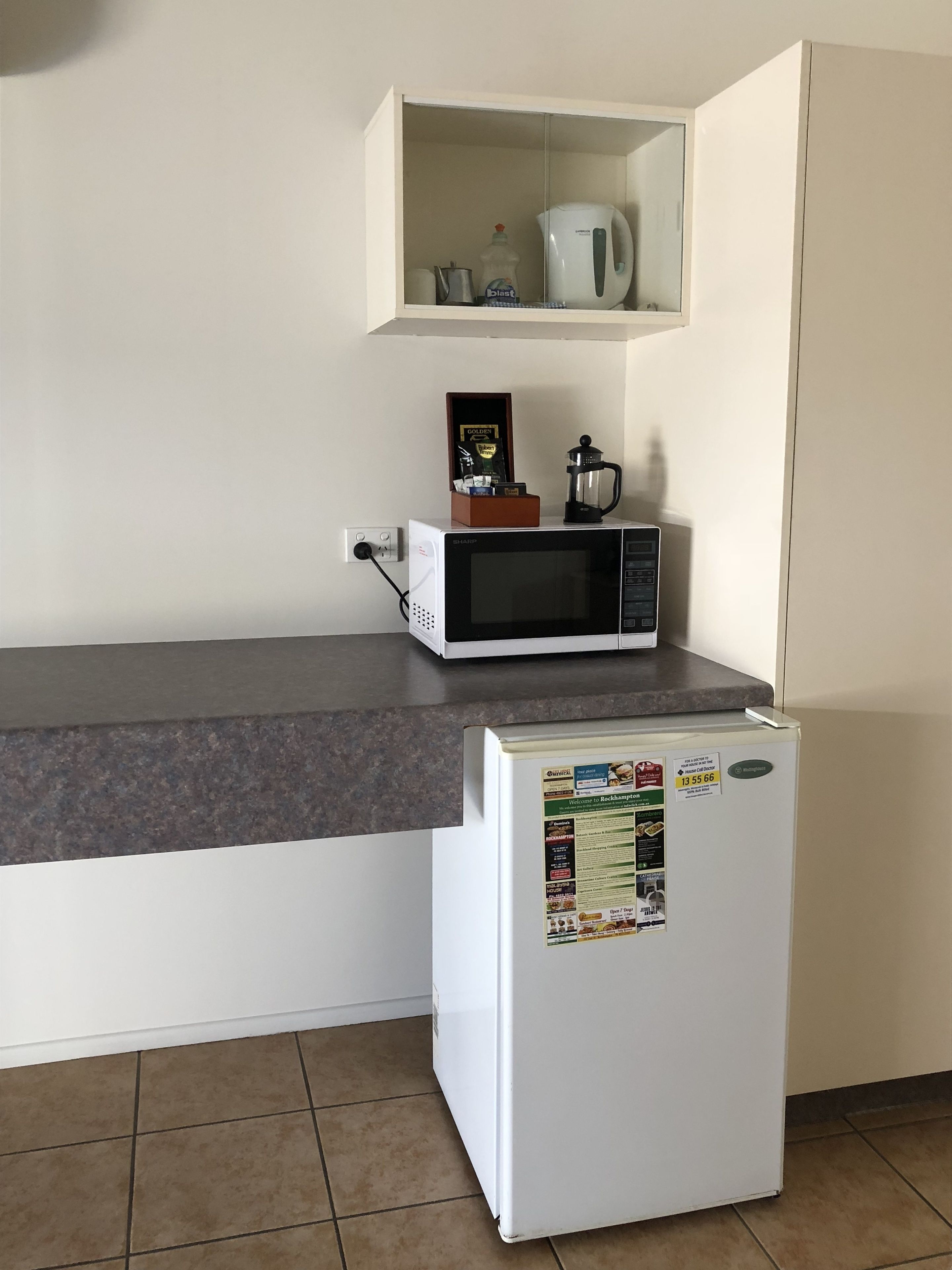 A compact kitchen with a microwave, a small refrigerator, and a shelf above the counter holding a coffee pot and a bottle.