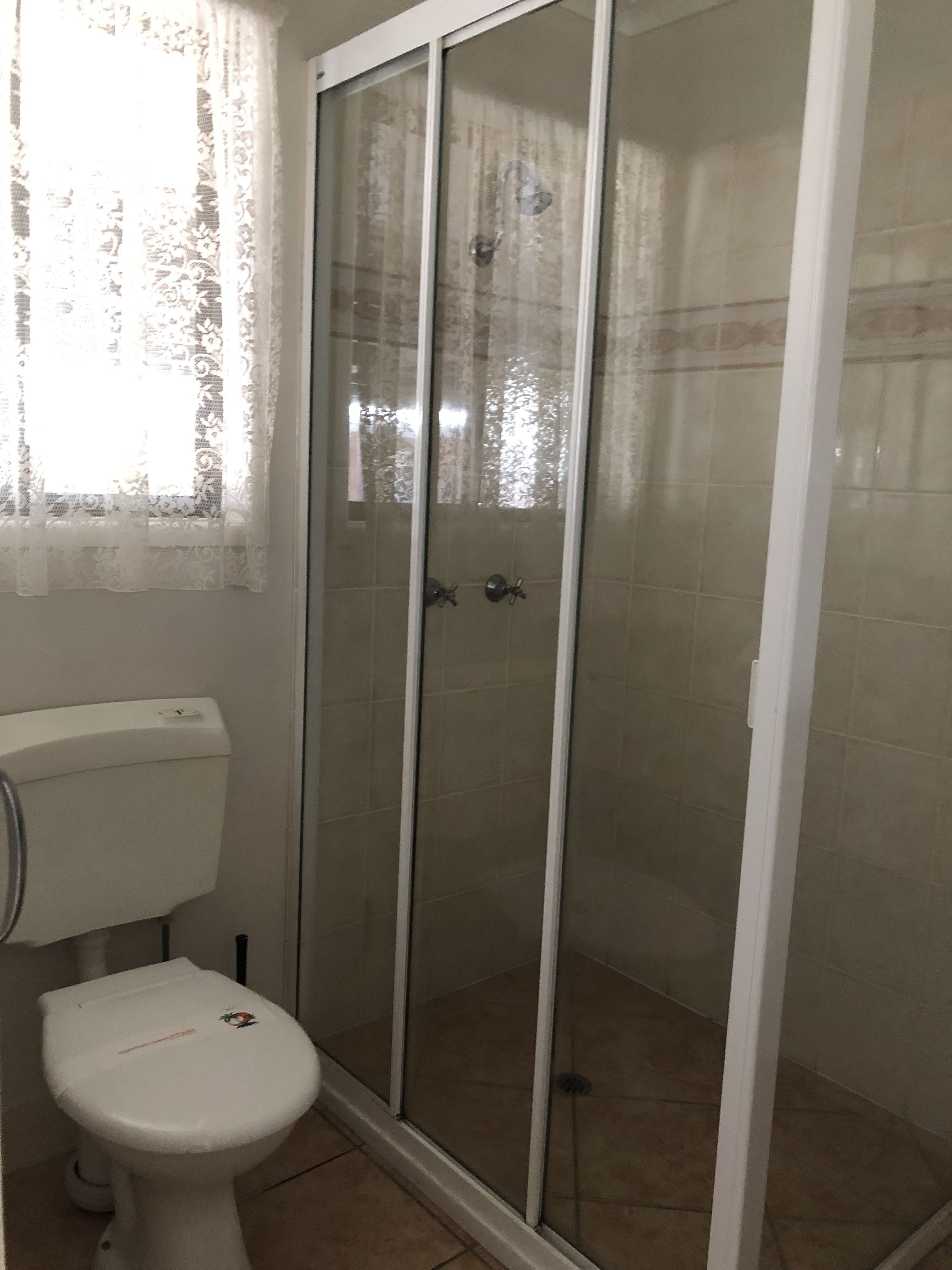 A bathroom with a toilet, a glass shower enclosure, and a window with lace curtains.
