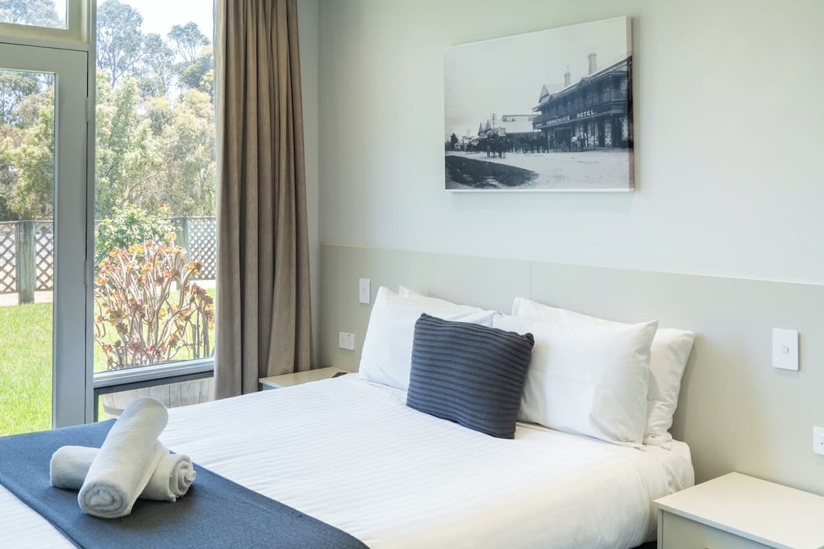 A hotel room with a bed, a nightstand, a ceiling fan, a window with curtains, and a wall-mounted picture.