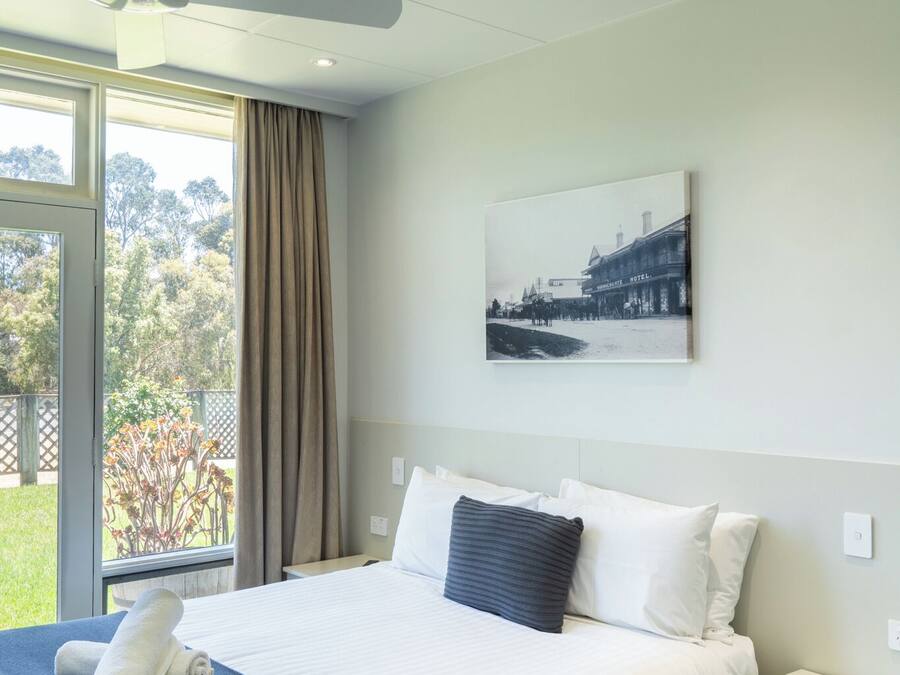 A hotel room with a bed, a nightstand, a ceiling fan, a window with curtains, and a wall-mounted picture.