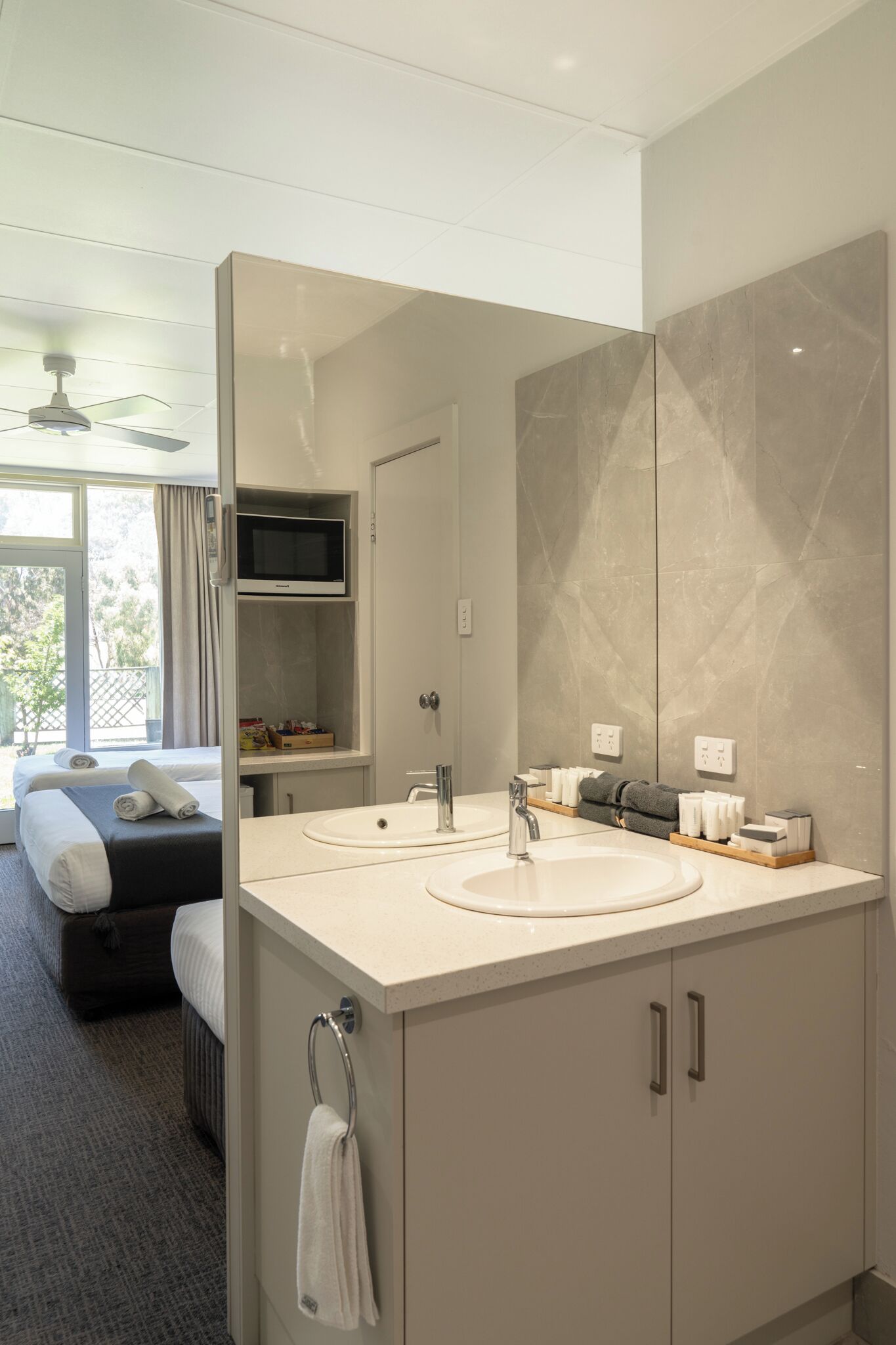 A modern hotel bathroom with a large mirror, two sinks, and a television above the mirror.