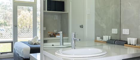 A modern bathroom with a large mirror, a sink, and a toilet. There is a towel rack with a towel hanging on it. The bathroom has a glass door leading to a balcony with a view of trees.