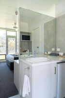 Un baño moderno con un espejo grande, un lavamanos y un inodoro. Hay un perchero con una toalla colgando. El baño tiene una puerta de cristal que da a un balcón con vistas a árboles.