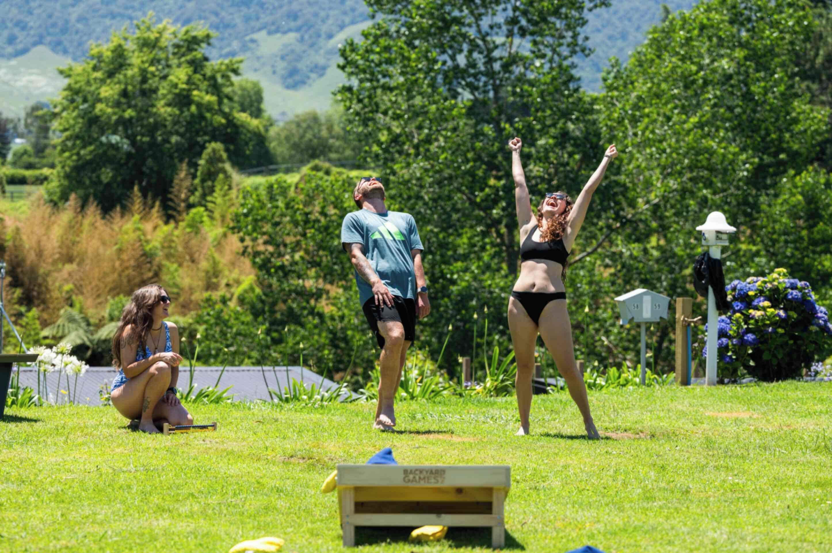Un espace extérieur recouvert d’herbe, avec une caisse en bois sur laquelle est inscrit « JEUX DE JARDIN », et une personne debout, les bras levés.