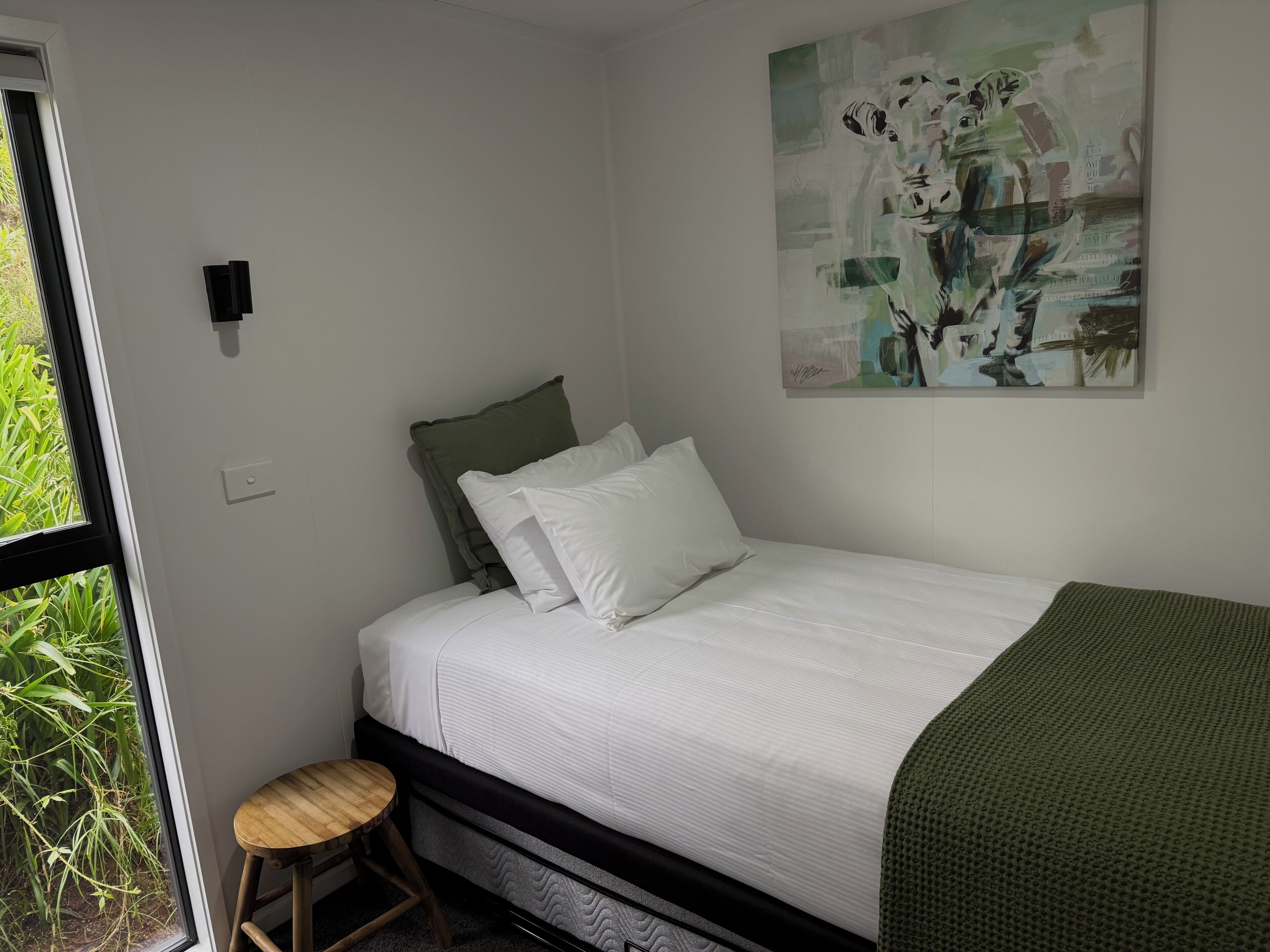 A bedroom with a bed, a green throw blanket, a small stool, and a painting on the wall.