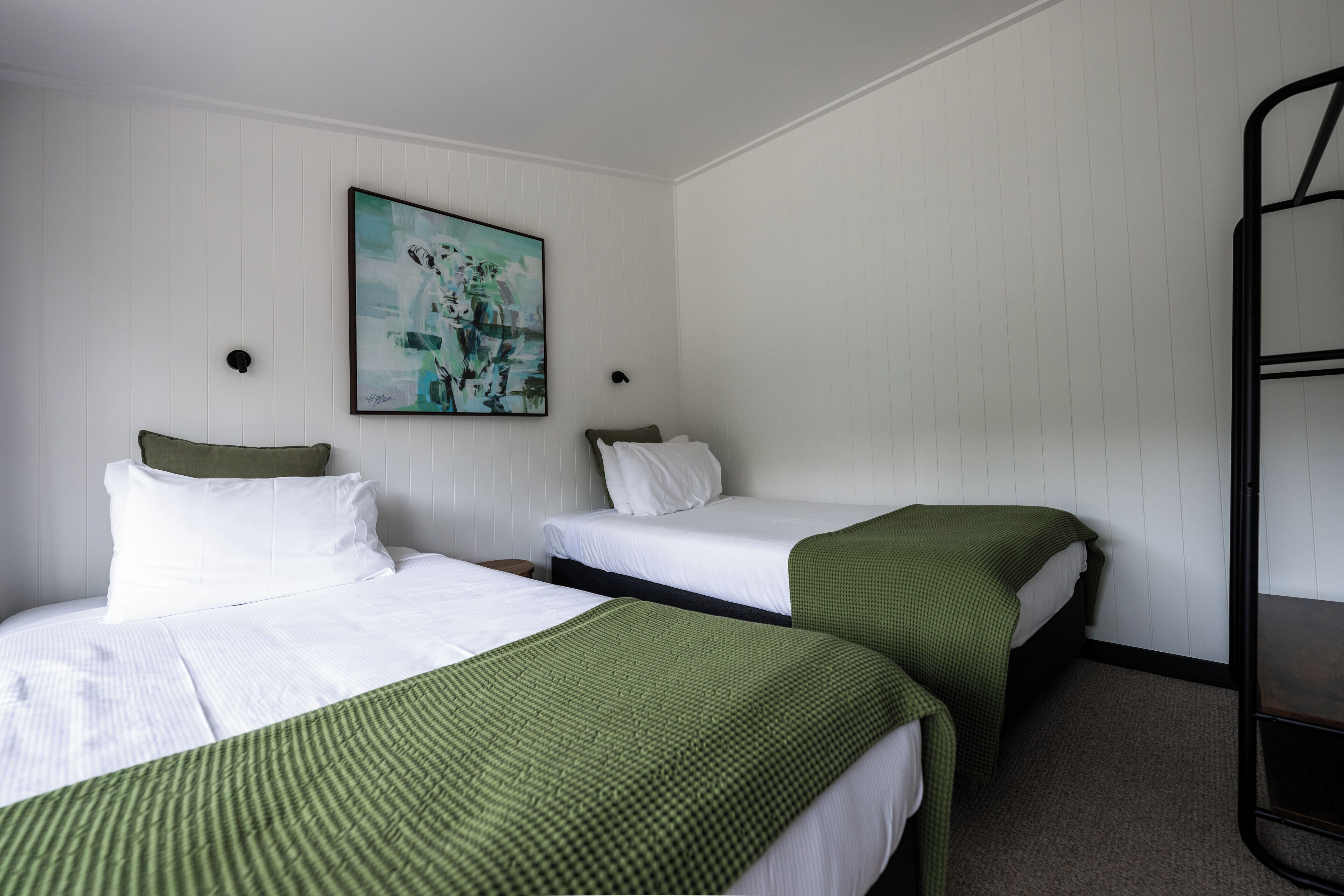 A room with two beds, white bedding, green throw blankets, a painting on the wall, and a metal shelf unit.
