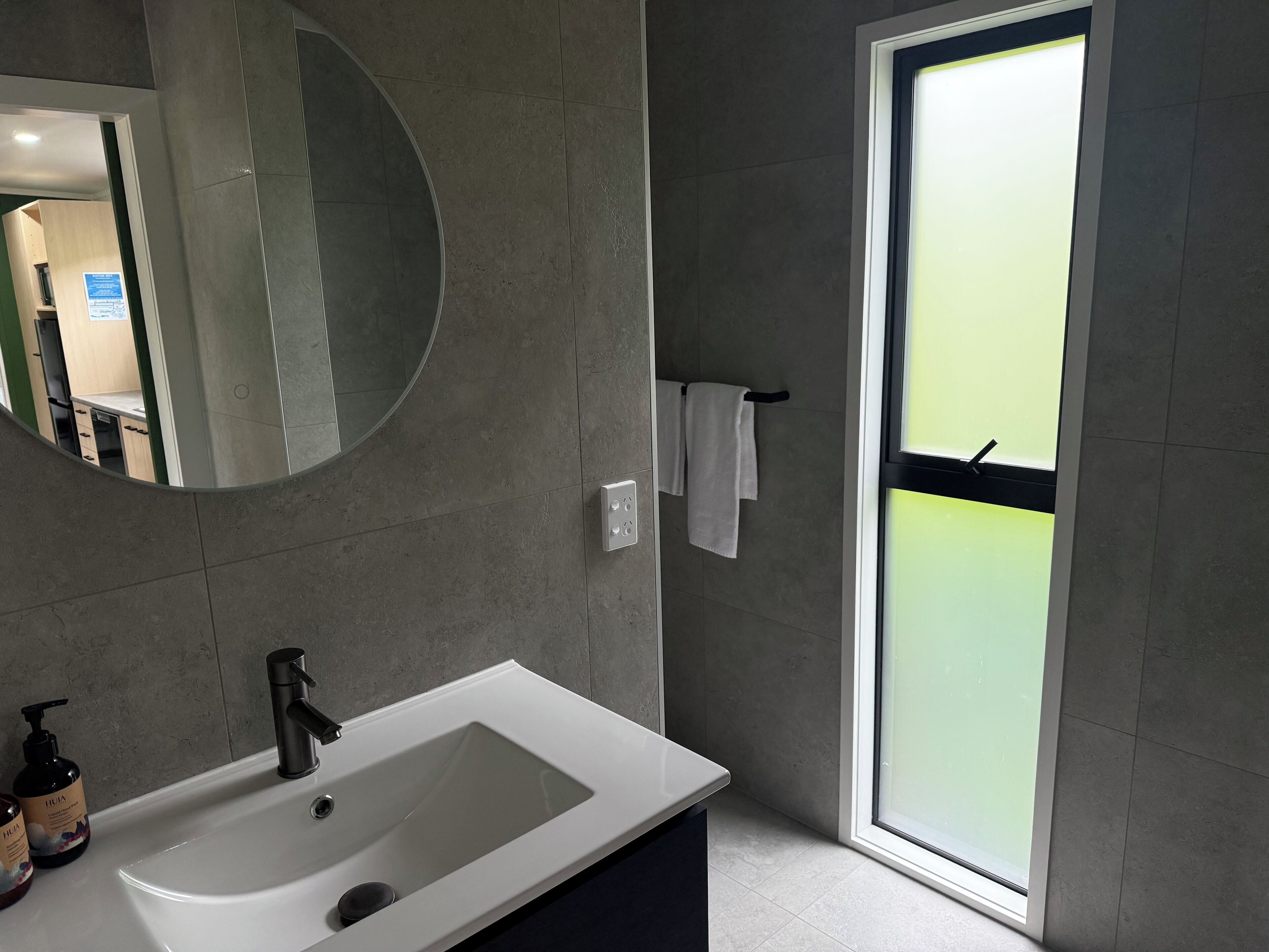 A modern bathroom with a white sink, a round mirror, and a glass door leading to a balcony.