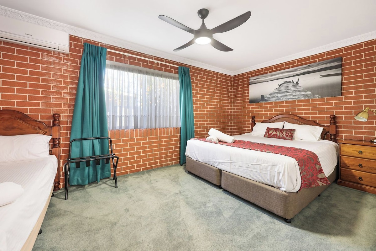 Une chambre d’hôtel avec un mur en briques, un lit avec une tête de lit en bois, une table de chevet, une chaise, un ventilateur de plafond et une fenêtre avec des rideaux.