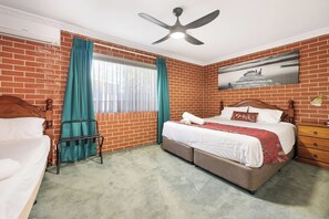 A hotel room with a brick wall, a bed with a wooden headboard, a bedside table, a chair, a ceiling fan, and a window with curtains. - Roseville Motel & Apartments (West Tamworth)