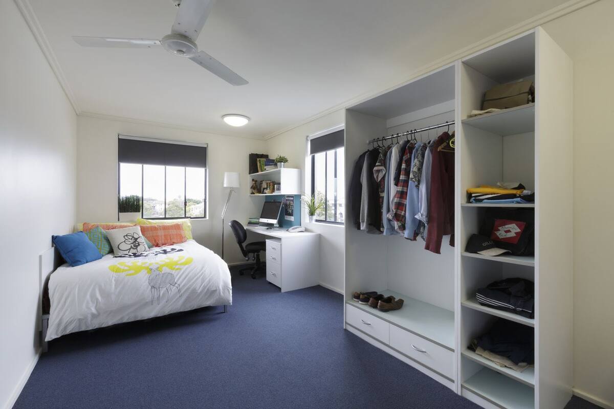 Quarto com cama, escrivaninha, guarda-roupa e ventoinha no teto.