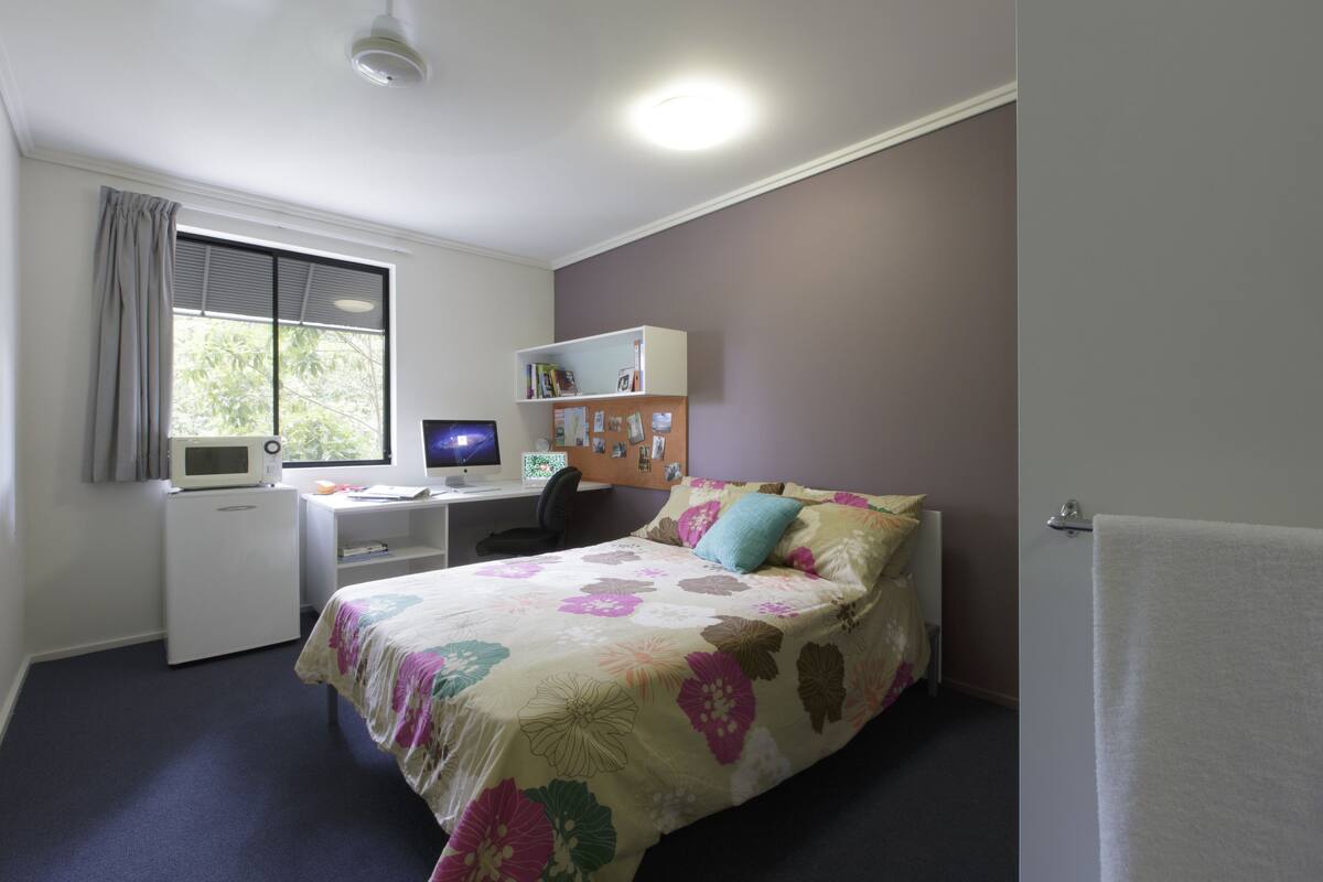 Um quarto com uma cama, uma escrivaninha com computador, estante de livros e janela com cortinas.