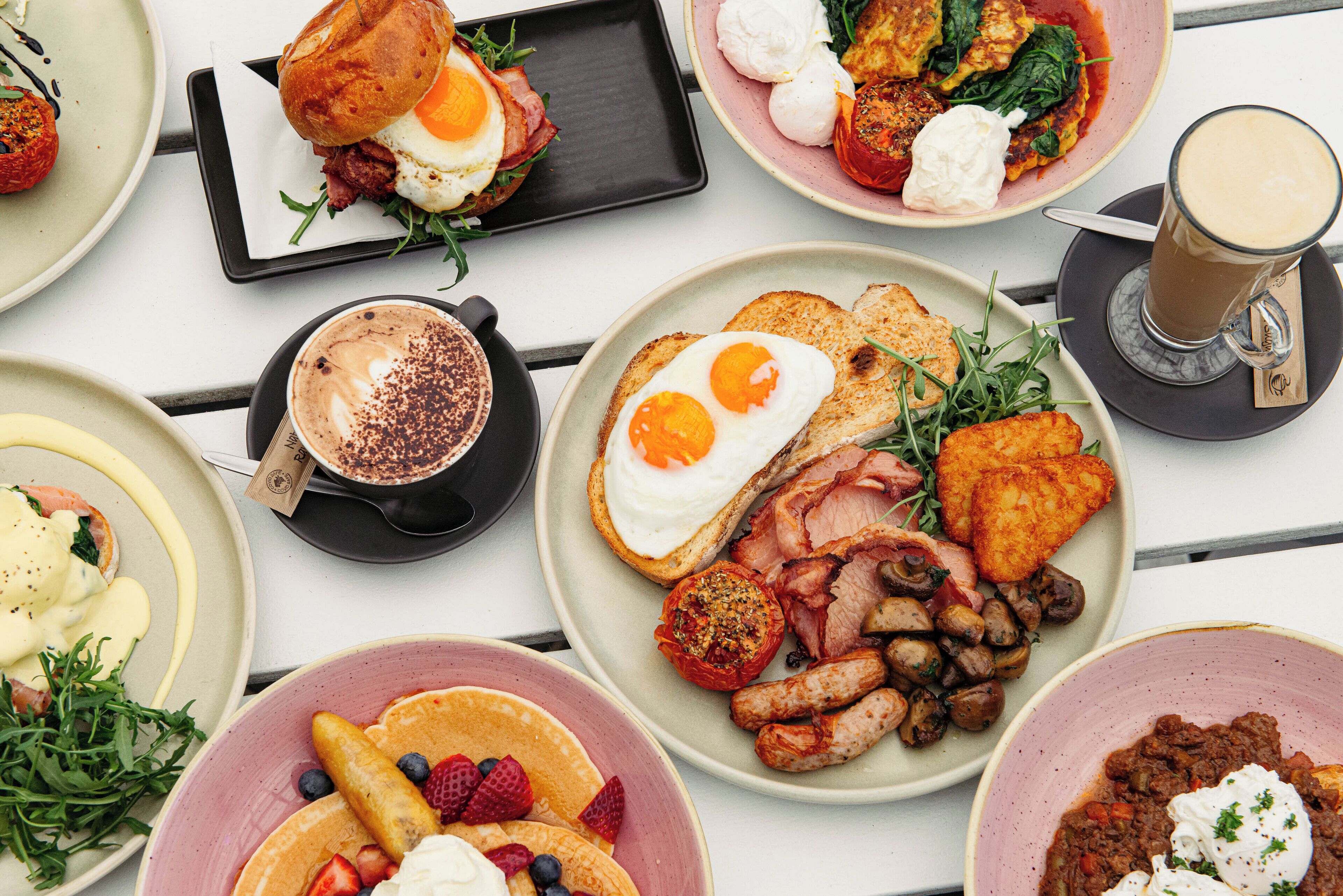 A variety of breakfast dishes including eggs, bacon, sausages, pancakes, and a coffee drink.