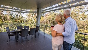 A covered outdoor area with a table and chairs, a glass railing, and a view of trees.