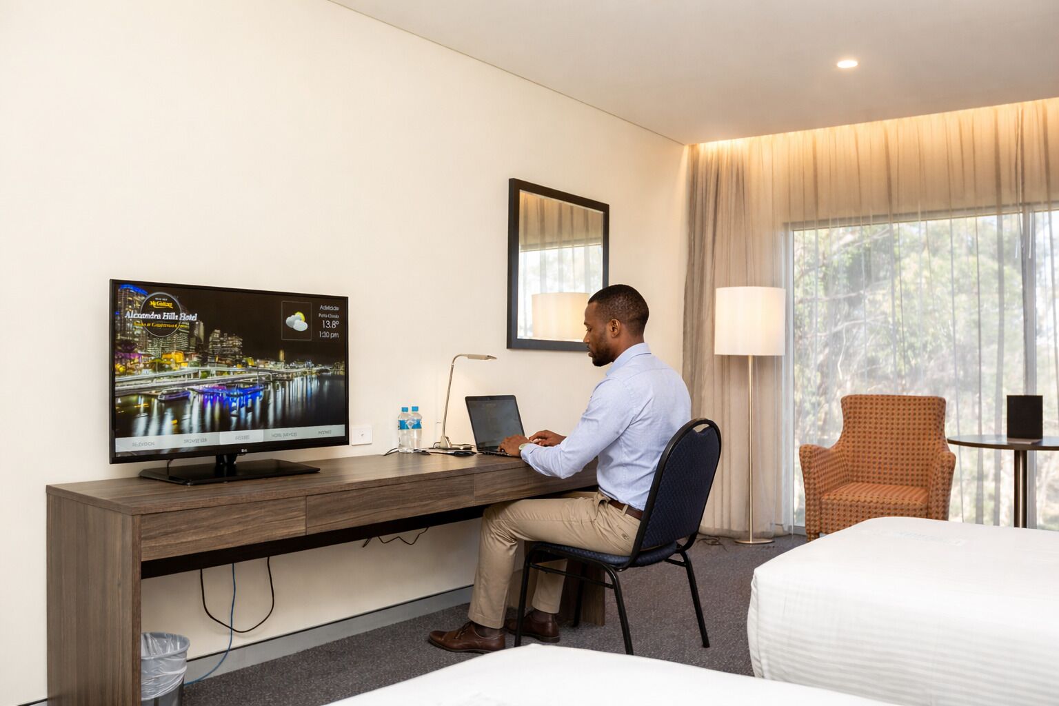 A hotel room with a bed, a desk with a laptop, a television displaying a cityscape, and a window with curtains.