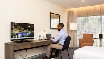 A hotel room with a bed, a desk with a laptop, a television displaying a cityscape, and a window with curtains.