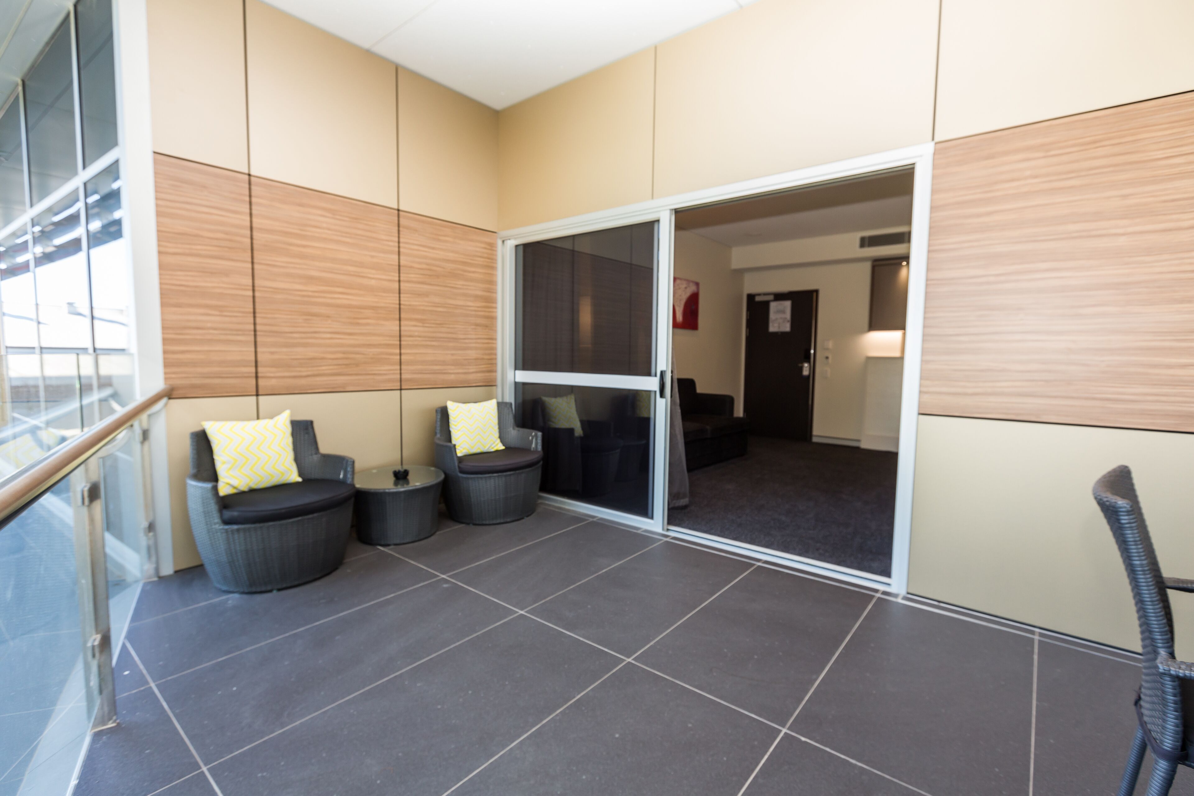 A modern indoor patio area with dark tiled flooring, two black wicker chairs with yellow pillows, and a glass door leading to another room.