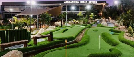 Un campo de mini golf bien iluminado con un edificio moderno al fondo que dice 'Alexandra Hills Suites'.