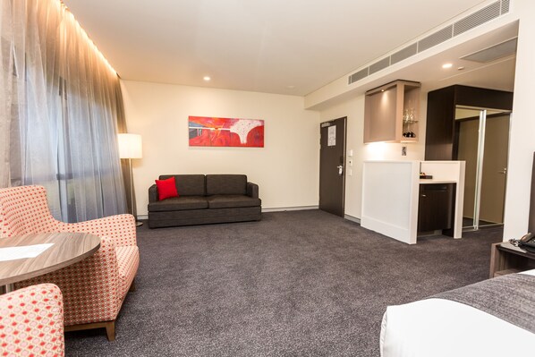 A hotel room with a grey carpet, a grey sofa with a red pillow, a patterned armchair, a small wooden table, a bed, a desk with a lamp, and a painting on the wall. - Alexandra Hills Hotel Suites (Alexandra Hills)