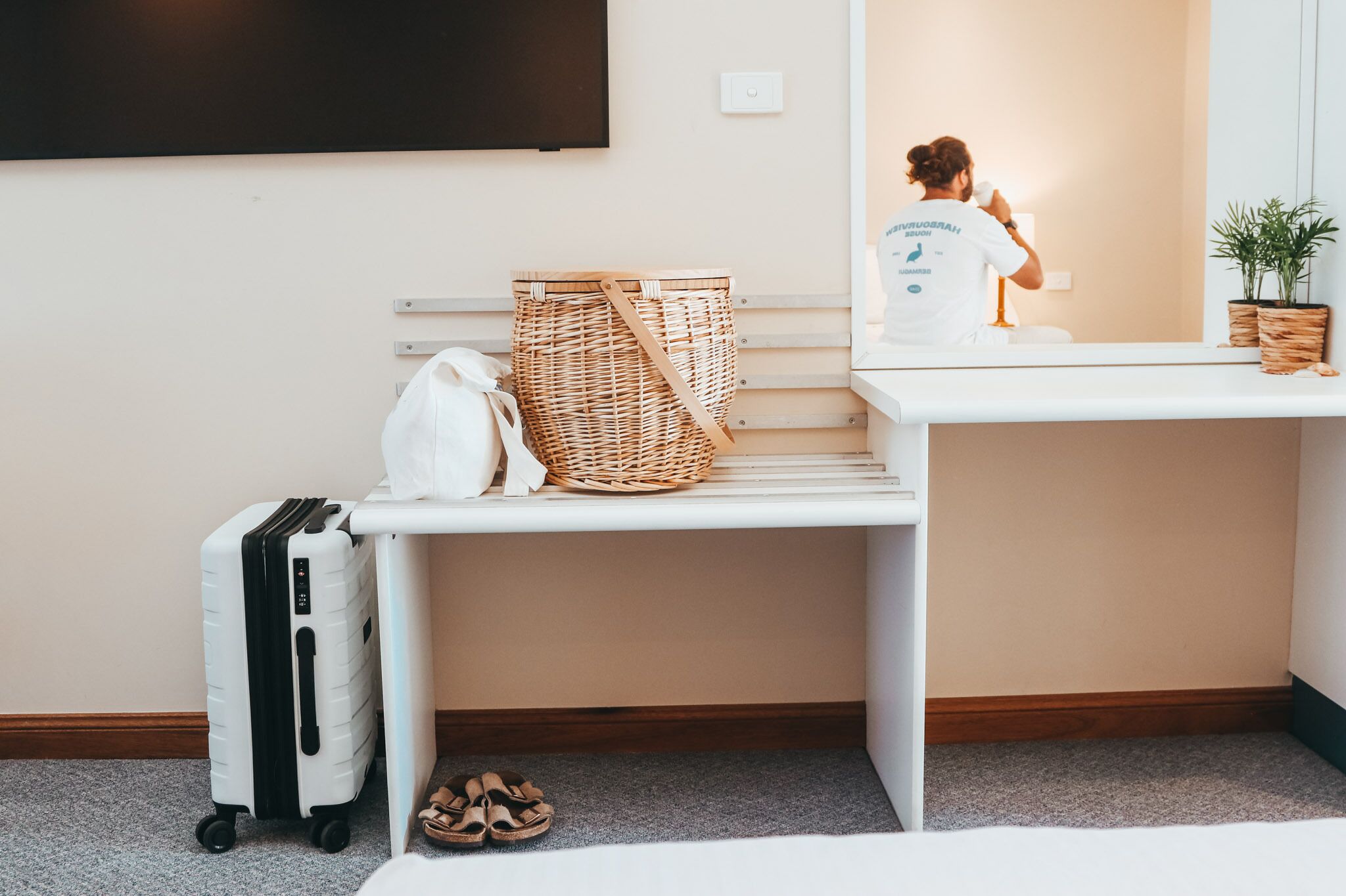 A hotel room with a white desk, a wicker basket, a white suitcase, and a pair of sandals on the floor.
