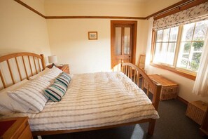 A bedroom with a wooden bed, a nightstand, a chair, a window with curtains, and a door. - Margate Cottage Boutique Bed & Breakfast (Margate)