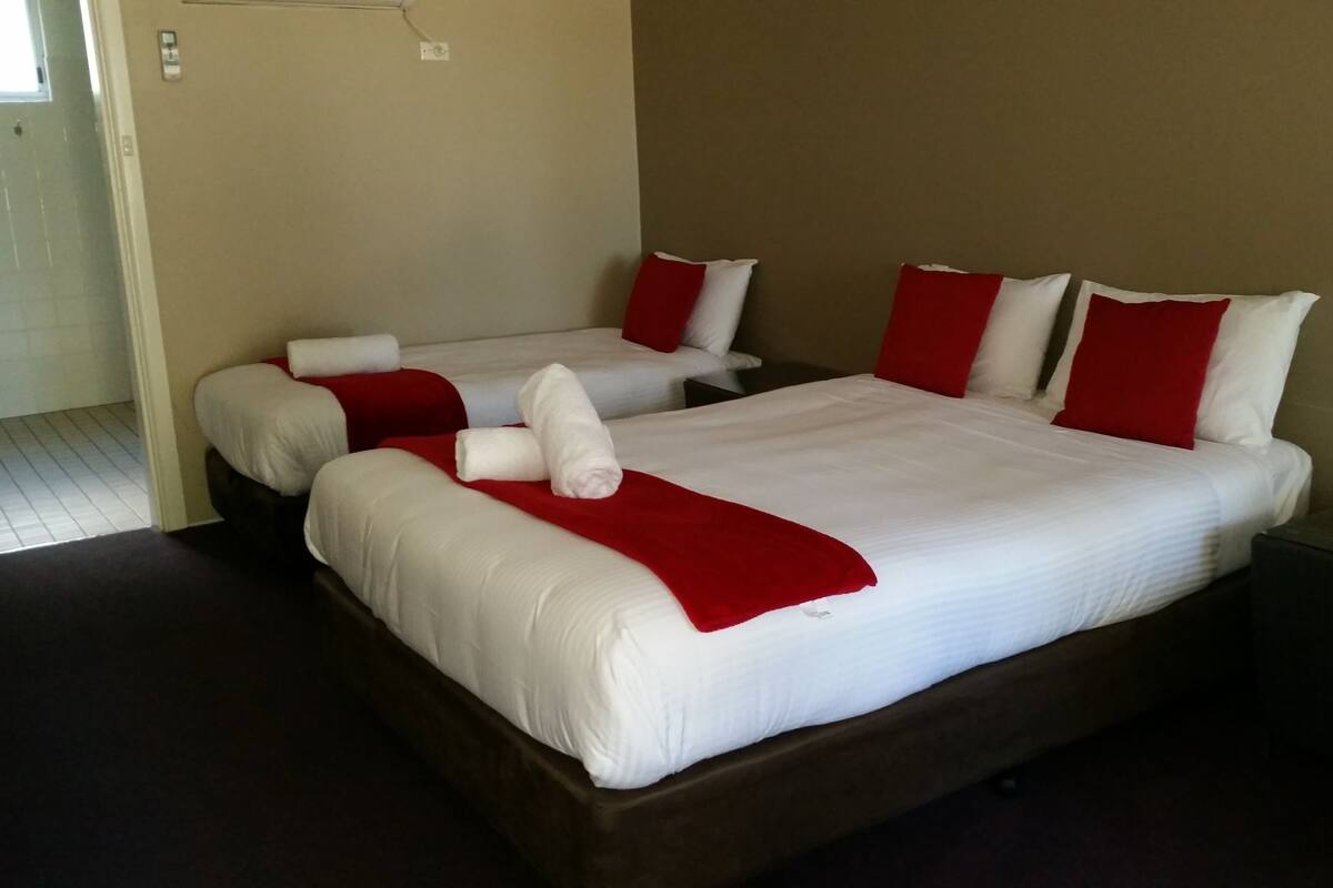 Ein Hotelzimmer mit zwei Betten, beide mit weißen Laken und roten Akzenten. Im Raum befindet sich ein kleiner Tisch und ein Stuhl.