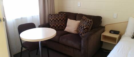 A small, furnished room with a brown couch, a round table, a chair, and a bedside table with a remote control.