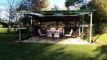 Een overdekt terras met een tafel en stoelen, omringd door een lattenhek en bomen.