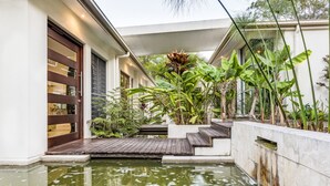 Un edificio moderno con terraza de madera, un pequeño estanque y plantas tropicales.