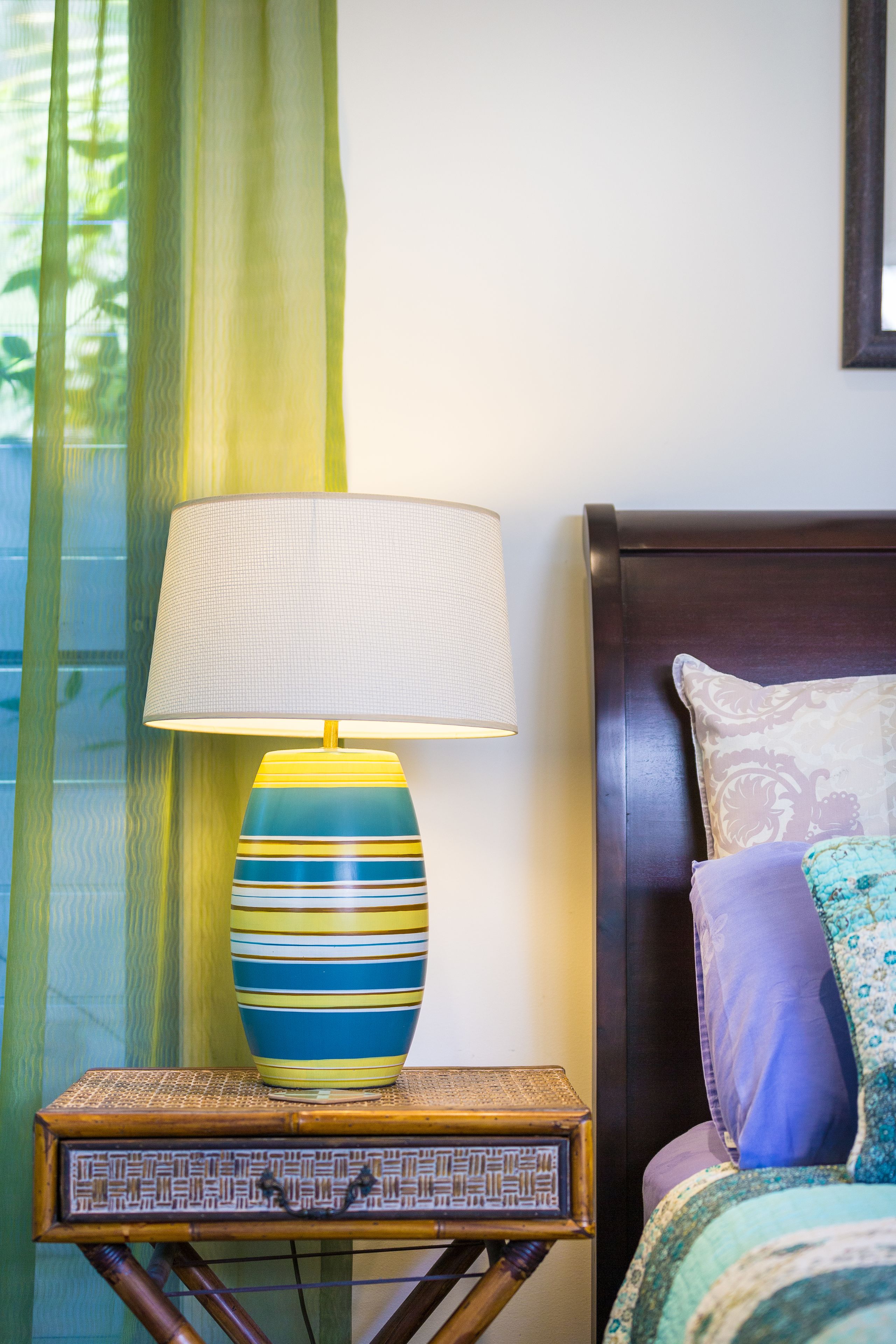 Un comodino con una lampada, un letto con cuscini decorati e una finestra con tende leggere.