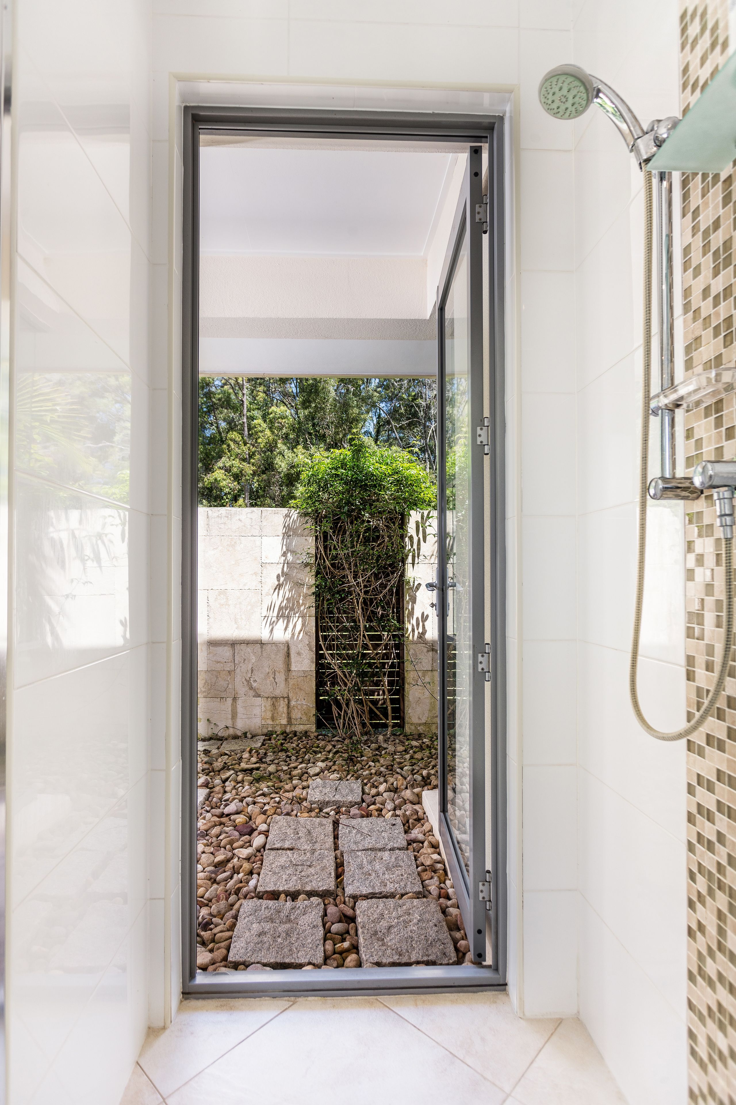 Un espace douche doté d’une porte en verre donnant accès à un jardin avec un chemin en pierre et de la verdure.