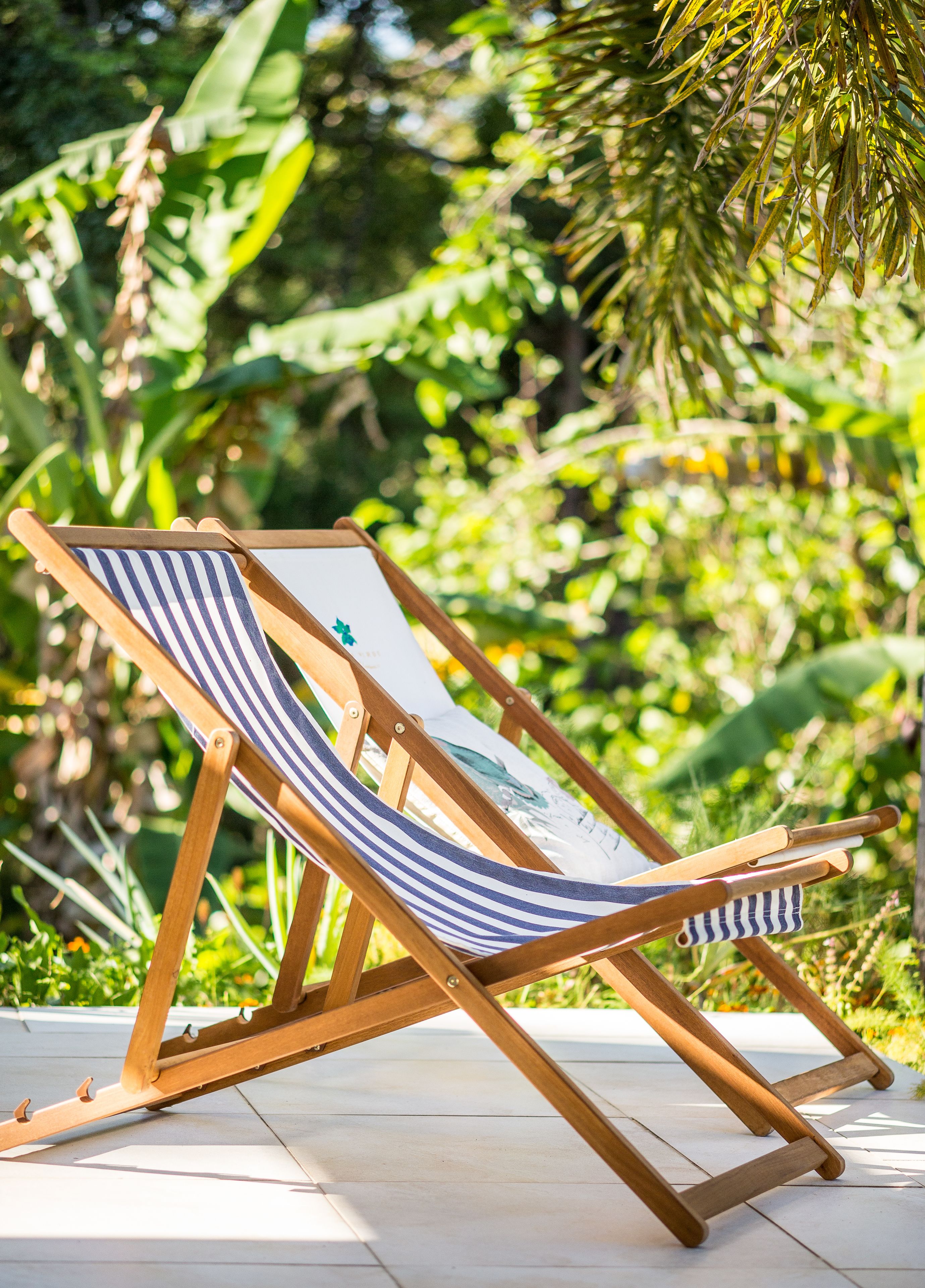 A wooden lounge chair with a striped cushion, positioned on a tiled patio with a lush green garden in the background.