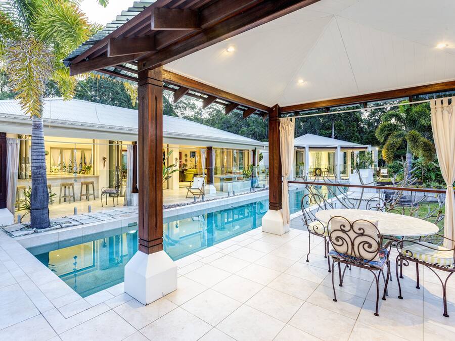 Ein überdachter Poolbereich mit Esstisch und Stühlen, einem Pool und Blick auf ein Haus mit Barbereich.