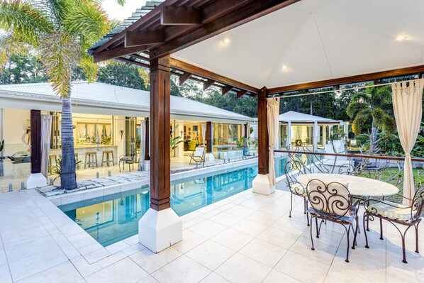 A covered outdoor pool area with a dining table and chairs, a pool, and a view of a house with a bar area.