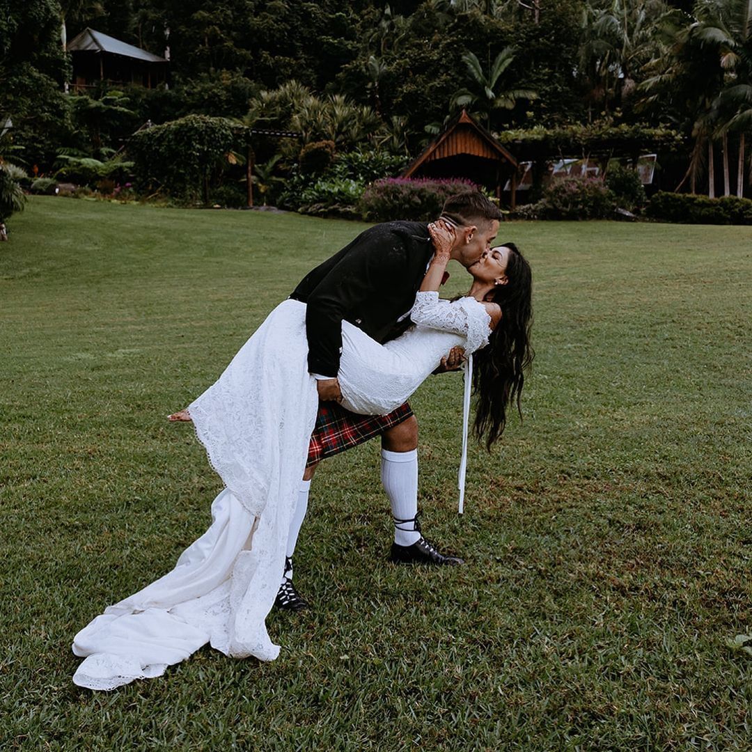 A couple embracing in a grassy outdoor area with a house and lush greenery in the background.