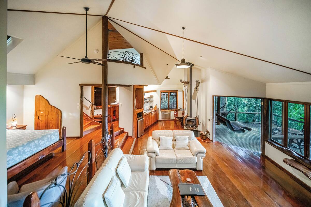 A spacious living area with a wooden floor, a sofa, a coffee table, and a dining table. There is a kitchen in the background and a large window with a view of the outdoors.